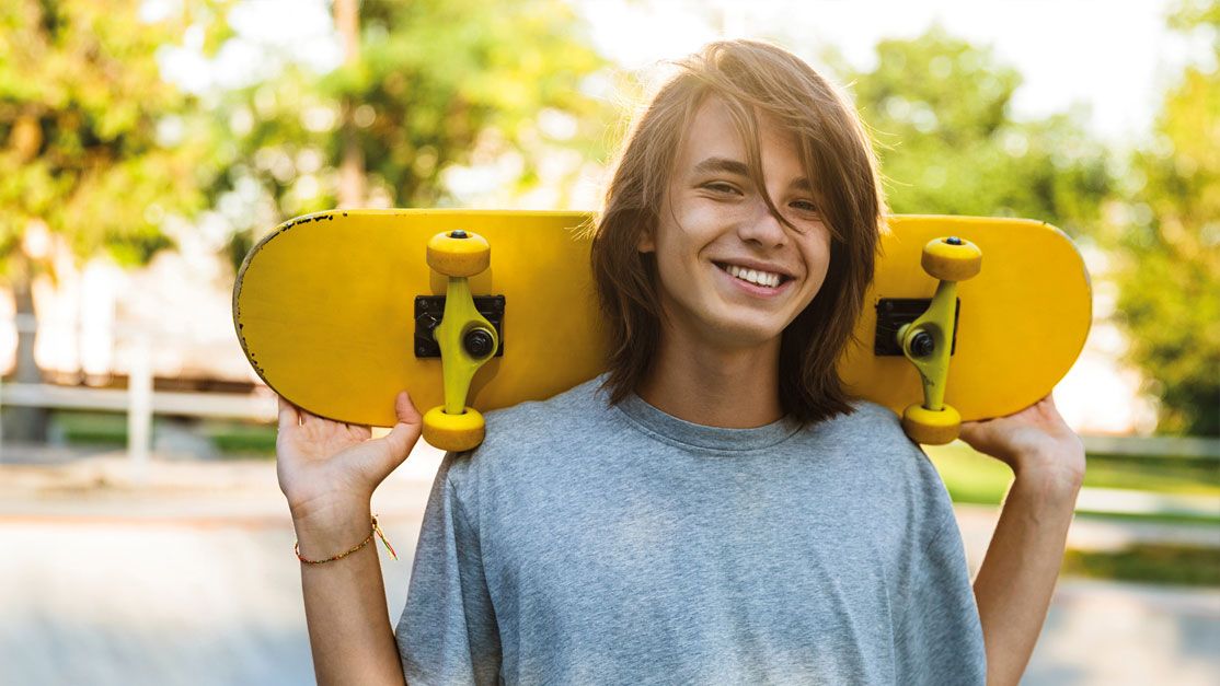 Ein Junge steht vor einer Halfpipe mit einem Skateboard auf den Schultern und lächelt glücklich.